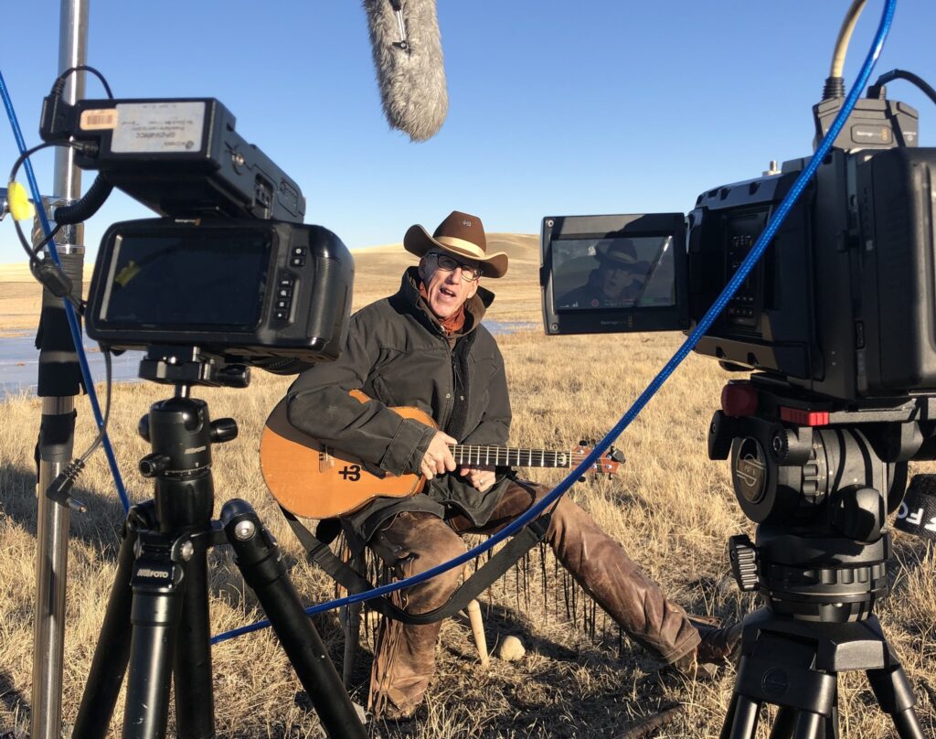 Wylie Gustafson performing with guitar on a Montana prairie during a Motion Noise shoot, framed between two Blackmagic cameras and a boom microphone